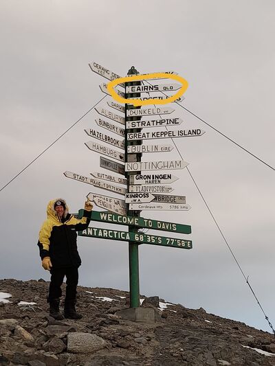 Jamie standing in front of a sign with directions to many cities around the world, pointing to Cairns