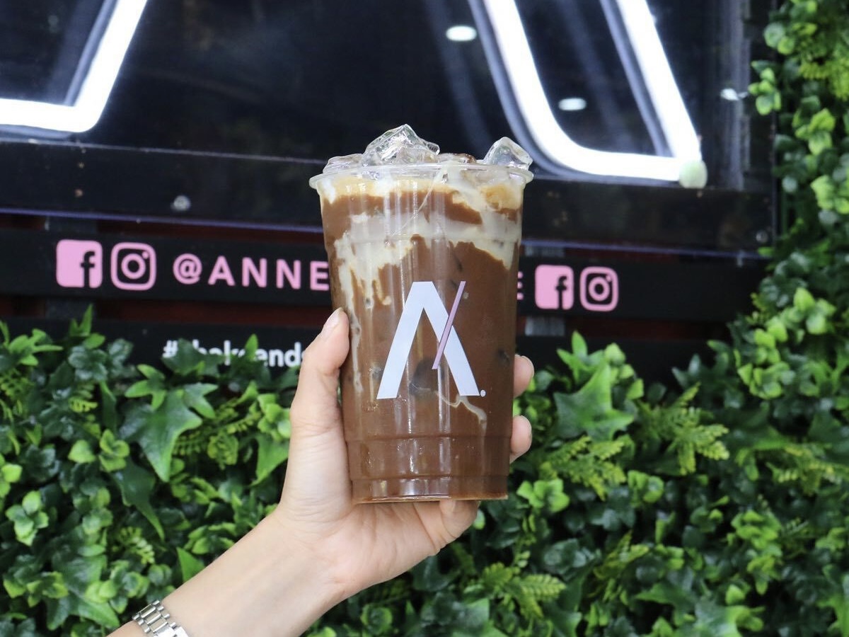 A hand holding up a Annee's Caphe Sua Da in front of a green wall